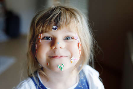 Little toddler girl playing with different colorful animal stickers. Concept of activity of children during pandemic virus quarantine. Happy funny child having fun with stick stickers on face.の写真素材