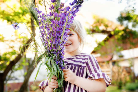 Cute adorable little toddler girl with huge bouquet of blossoming violet lupnies flowers. Portrait of smiling preschool child in domestic garden on warm spring or summer day. Summertimeの写真素材