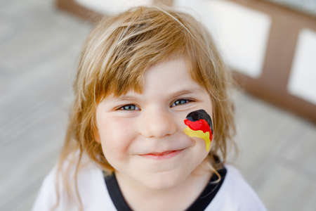 Little blond preschool girl watching soccer football cup game on public viewing. Happy joyful excited child about winning game match of favorite national football team. girl with German flag on face.の写真素材