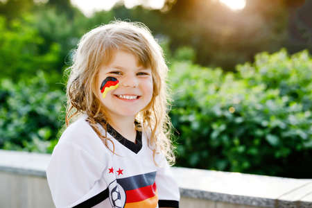 Little blond preschool girl watching soccer football cup game on public viewing. Happy joyful excited child about winning game match of favorite national football team. girl with German flag on face.の写真素材