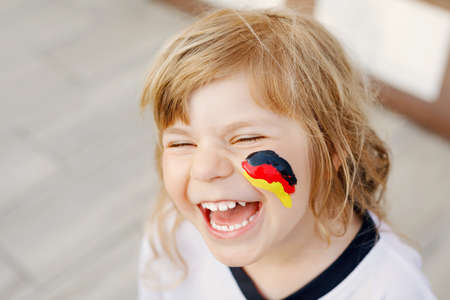 Little blond preschool girl watching soccer football cup game on public viewing. Happy joyful excited child about winning game match of favorite national football team. girl with German flag on face.の写真素材
