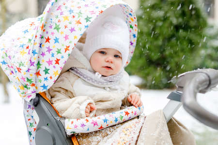 Cute little beautiful baby girl sitting in the pram or stroller on cold snowy winter day. Happy smiling child in warm clothes, fashion stylish baby coat. Babys first snow. Winter walk outdoors.の写真素材