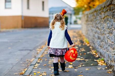 Little toddler girl dressed as a witch trick or treating on Halloween. Happy child outdoors, with orange funny hat and pumpkin bag for sweet haunt. Family festival season in october. Outdoor activityの写真素材