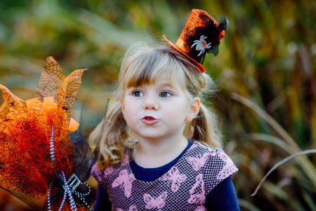 Portrait of little toddler girl dressed as a witch celebrates Halloween. Happy child outdoors, with...