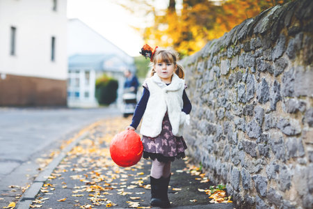 Little toddler girl dressed as a witch trick or treating on Halloween. Happy child outdoors, with orange funny hat and pumpkin bag for sweet haunt. Family festival season in october. Outdoor activityの写真素材