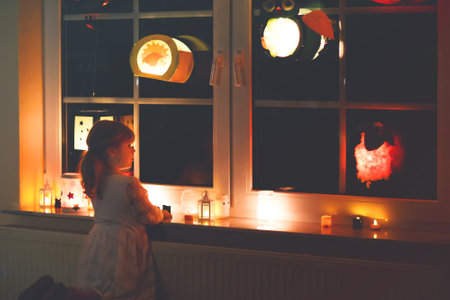 Little kid girl sitting by window with selfmade hand crafted lanterns with candles for St. Martin procession. Toddler child looking at glowing lantern. German tradition Martinsumzug. Home decorationの写真素材