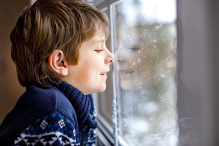 Happy adorable kid boy sitting near window and looking outside on snow on Christmas day or morning. Smiling child fascinated with snowfall and big snowflakesの写真素材