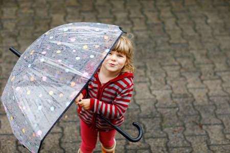Little toddler girl playing with big umbrella on rainy day. Happy positive child running through rain, puddles. Preschool kid with rain clothes and rubber boots. Children activity on bad weather day.の写真素材