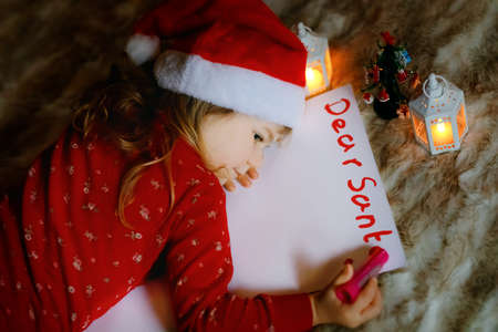 Little cute toddler girl in bed under Christmas tree, writing wish list letter to Santa Claus at home, indoors. Traditional Christian festival. Happy kid child waiting for gifts on xmas.の写真素材