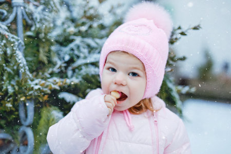 Portrait of little toddler girl walking outdoors in winter. Cute toddler eating sweet lollypop candy. Child having fun on cold snow day. Wearing warm baby pink clothes and hat with bobbles.の写真素材