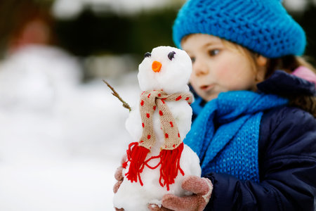 Cute little toddler girl making mini snowman and eating carrot nose. Adorable healthy happy child playing and having fun with snow, outdoors on cold day. Active leisure with children in winterの写真素材
