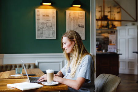 Portrait of young woman using technology of smartphone and looking at laptop. Young woman freelancer using laptop computer working in restaurant or hipster indoor cafe with coffee. Home office conceptの写真素材