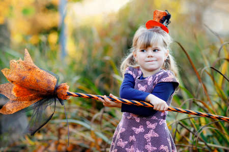 Cute little toddler girl dressed as a witch celebrates Halloween. Happy child outdoors, with orange funny hat and hold witch broom. Beautiful family festival season in october. Outdoor activityの写真素材