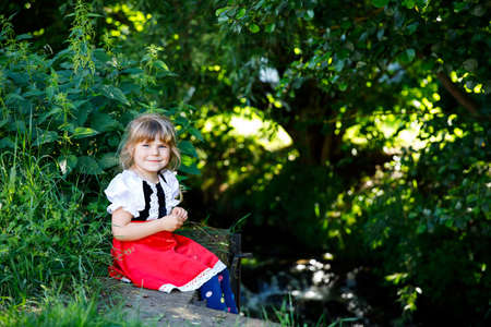 Little preschool girl in red riding hood dress sitting in forest by a brook. Cute toddler child enjoying walk in nature on warm summer day. Activity with children outdoors.の写真素材