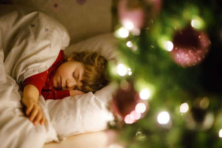 Little cute toddler girl sleeping under Christmas tree and dreaming of Santa at home, indoors. Traditional Christian festival. Happy kid child waiting for gifts on xmas. Cozy soft lightの写真素材