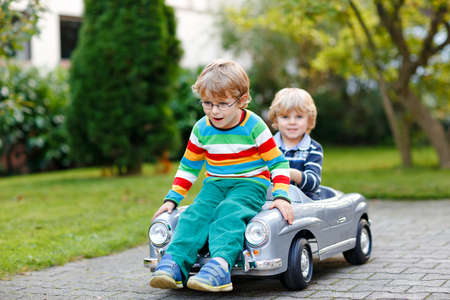 Two little preschool boys playing with big old toy car in summer garden, outdoors. Happy children play together, driving car. Outdoor activity for kids. Siblings and friends on warm dayの写真素材