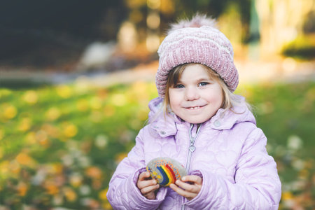 Adorable little toddler girl with painted rainbow on stone during pandemic quarantine, outdoors....