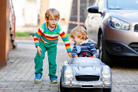 Two little preschool boys playing with big old toy car in summer garden, outdoors. Happy children play together, driving car. Outdoor activity for kids. Siblings and friends on warm dayの写真素材