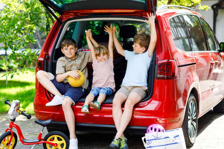 Three children, two boys and preschool girl sitting in car trunk before leaving for summer vacation with parents. Happy kids, siblings, brothers and sister with suitcases and toys going on journeyの写真素材