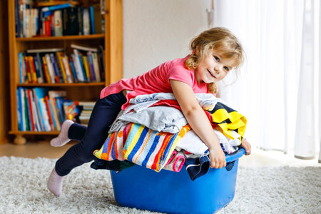 Little girl with a big basket of fresh clean laundry ready for ironing. Happy beautiful toddler and baby daughter helping mother with housework and clothes.の写真素材