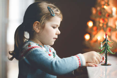 Litte toddler girl sitting by window and decorating small glass Christmas tree with tiny xmas toys. Happy healthy child celebrate family traditional holiday. Adorable baby.の写真素材