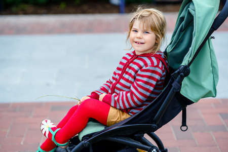 Portrait of little cute toddler girl sitting in stroller or pram and going for a walk. Happy cute baby child having fun outdoors. Healthy daughter. Street traffic in the city.の写真素材