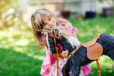 Little preschool girl hugging with rocking horse toy. Happy child in princess dress on sunny summer day in garden. Girl in love with her favourite old vintage toy animal.の写真素材