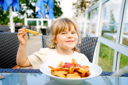 Portrait of happy smiling preschool girl eating french fries with tomato ketchup in restaurant on...