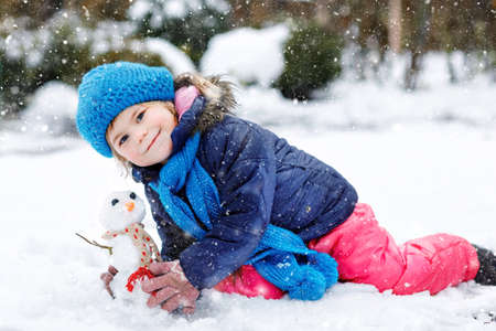 Cute little toddler girl making mini snowman and eating carrot nose. Adorable healthy happy child playing and having fun with snow, outdoors on cold day. Active leisure with children in winterの写真素材