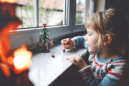 Litte toddler girl sitting by window and decorating small glass Christmas tree with tiny xmas toys. Happy healthy child celebrate family traditional holiday. Adorable baby.の写真素材