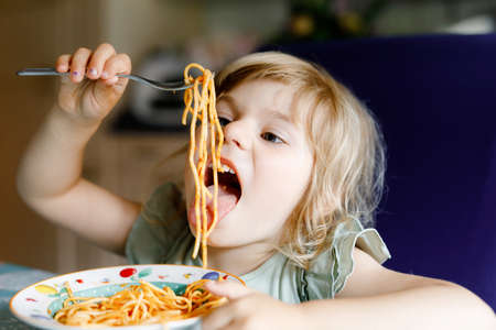 Adorable toddler girl eat pasta spaghetti with tomato bolognese with minced meat. Happy preschool child eating fresh cooked healthy meal with noodles and vegetables at home, indoors.の写真素材