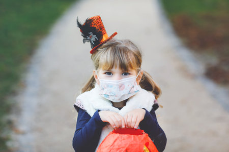Little toddler girl dressed as a witch with medical mask on face trick or treating on Halloween. Child outdoors, with funny hat and bag for sweet haunt. Pandemic corona virus quarantine lockdown timeの写真素材