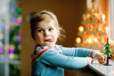 Litte toddler girl sitting by window and decorating small glass Christmas tree with tiny xmas toys....