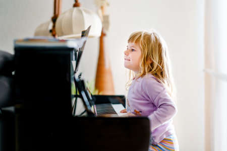 Beautiful little toddler girl playing piano in living room. Cute preschool child having fun with learning to play music instrument. Early musical education for children.の写真素材