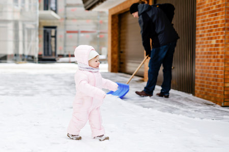 Adorable little baby girl making first steps outdoors in winter through snow. Cute toddler child learning walking. On background father, young man cleaning street with snow shovel. Happy familyの写真素材
