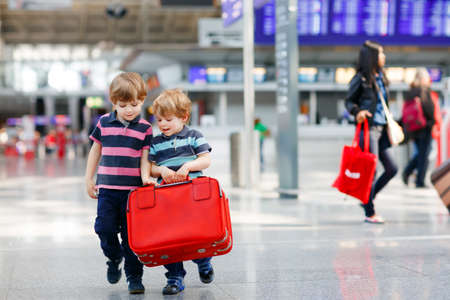 Two little sibling boys having fun and going on vacations trip with suitcase at airport, indoors. Happy twins brothers walking to check-in or boarding. Family on flight, journey on planeの写真素材
