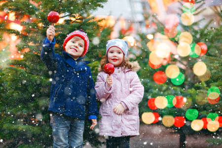 Two little smiling kids, preschool boy and girl eat sweet sugared apple on German Christmas market. Happy siblings children with lights on background and xmas trees. Family funny brother and sister.の写真素材