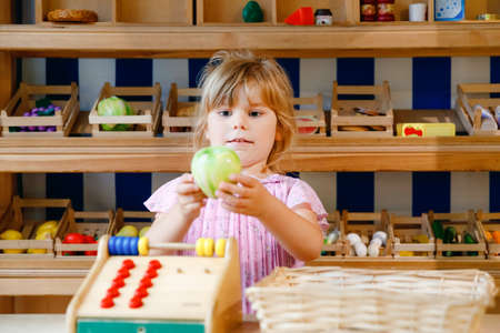 Little preschool girl play with food and grocery wooden toys. Happy active child playing role game as cashier or seller, in wood shop or supermarket. Education, activity for kids.の写真素材