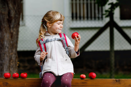 Little cute girl eat red ripe apple. Happy smiling preschool child with satchel holding fresh bio fruits. Apples from harvest.の写真素材