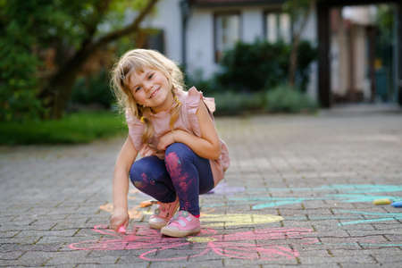 Little preschool girl painting with colorful chalks flowers on ground on backyard. Positive happy toddler child drawing and creating pictures on asphalt. Creative outdoors children activity in summer.の写真素材