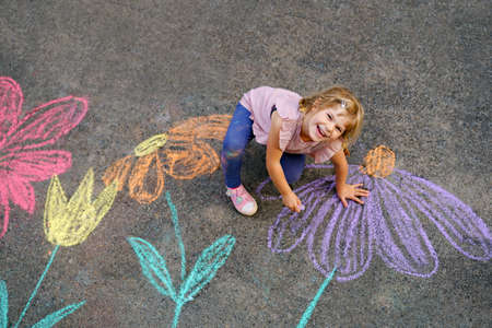 Little preschool girl painting with colorful chalks flowers on ground on backyard. Positive happy toddler child drawing and creating pictures on asphalt. Creative outdoors children activity in summer.の写真素材