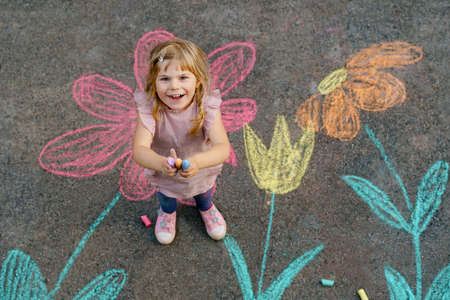 Little preschool girl painting with colorful chalks flowers on ground on backyard. Positive happy toddler child drawing and creating pictures on asphalt. Creative outdoors children activity in summer.の写真素材