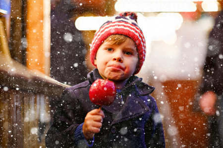 Cute little smiling preschool boy on German Christmas market. Happy child in winter clothes eating sweet sugared glazed xmas apple on with lights on background. Family, tradition, celebration conceptの写真素材
