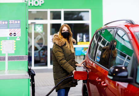 Woman wear medical mask at self-service gas station, hold fuel nozzle, refuel the car with petrol during corona virus pandemic lockdown. People in masks as preventive measure and covid protectionの写真素材