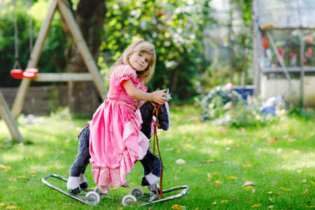 Little preschool girl hugging with rocking horse toy. Happy child in princess dress on sunny summer day in garden. Girl in love with her favourite old vintage toy animal.の写真素材