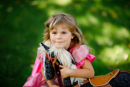 Little preschool girl hugging with rocking horse toy. Happy child in princess dress on sunny summer day in garden. Girl in love with her favourite old vintage toy animal.の写真素材