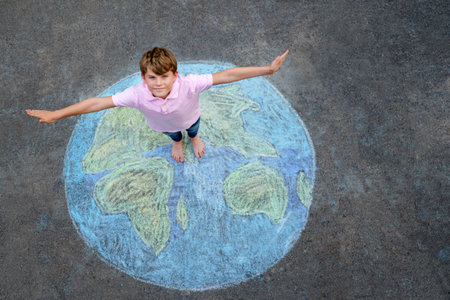 Little school boy with earth globe painting with colorful chalks on ground. Positive kid child. Happy earth day concept. Creation of children for saving world, environment and ecology.の写真素材