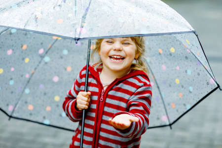 Little toddler girl playing with big umbrella on rainy day. Happy positive child running through rain, puddles. Preschool kid with rain clothes and rubber boots. Children activity on bad weather day.の写真素材