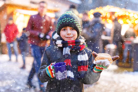 Little cute kid boy eating German sausage and drinking hot children punch on Christmas market. Happy child on traditional family market in Germany. Laughing boy in colorful winter clothesの写真素材