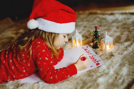 Little cute preschool girl in pajamas under Christmas tree, writing wish list letter to Santa Claus...